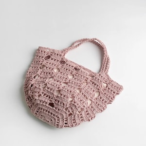 Puede incluir: Un bolso tote de ganchillo rosa polvoriento con dos asas. El bolso presenta un diseño de tejido abierto y un borde festoneado. El bolso está hecho de un material suave y flexible.
