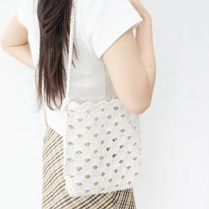 Puede incluir: Un bolso de crochet blanco con un borde festoneado y una larga correa trenzada. El bolso presenta un diseño geométrico abierto. El bolso se lleva al hombro. El fondo es una pared blanca lisa.