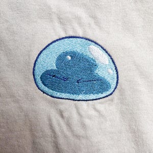 Könnte beinhalten: Gestickter Aufnäher mit einem blauen Schleim-Charakter in einer hellblauen Blase mit dunkelblauem Umriss. Der Aufnäher befindet sich auf einem hellgrauen Stoffhintergrund.