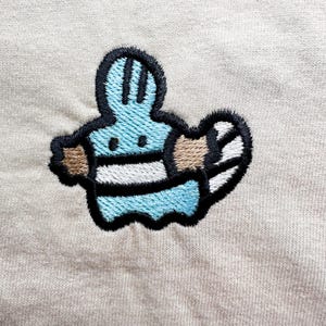 May include: Embroidered patch featuring a cartoon character in light blue, white, and brown, outlined in black. The design is on a beige fabric background. The character has a striped body and a rounded shape.