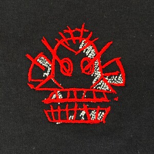 May include: Black fabric with a red embroidered skull design. The skull features a crown-like structure and a toothy grin. The embroidery has a textured appearance with white accents, creating a striking contrast against the dark background.
