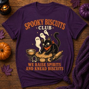 Pode incluir: Uma t-shirt roxa com um gráfico de um gato preto com uma bandana laranja, a amassar massa com pequenos fantasmas por cima. O texto diz SPOOKY BISCUITS CLUB e WE RAISE SPIRITS AND KNEAD BISCUITS, acompanhado por uma taça, um rolo e cortadores de bolachas.