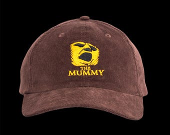 May include: Brown corduroy baseball cap with a yellow embroidered graphic of a mummy's face and the text "THE MUMMY" in yellow. The cap has a curved brim and a rounded crown, suitable for casual wear.