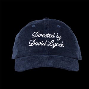 May include: Navy blue corduroy baseball cap with the embroidered text "Directed by David Lynch" in white script. The cap has a curved brim and a rounded crown. The background is black.