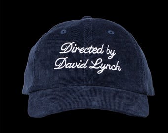 May include: Navy blue corduroy baseball cap with the embroidered text "Directed by David Lynch" in white script. The cap has a curved brim and a rounded crown. The background is black.