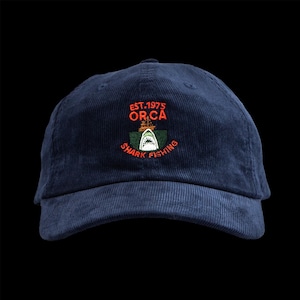 May include: Navy blue corduroy baseball cap with an embroidered design. The design features a shark, boat, and the text "EST.1975 OR.CA SHARK FISHING" in orange and white. The cap has a curved brim and a top button.