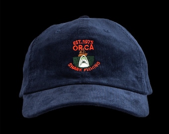 May include: Navy blue corduroy baseball cap with an embroidered design. The design features a shark, boat, and the text "EST.1975 OR.CA SHARK FISHING" in orange and white. The cap has a curved brim and a top button.