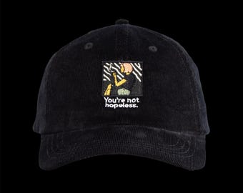 May include: A black corduroy baseball cap featuring an embroidered square patch on the front. The patch depicts a stylized figure in yellow and pink against a black and white striped background, with the text You're not hopeless. embroidered below.