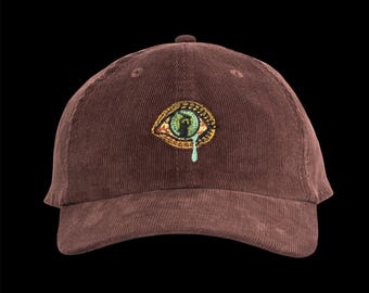 May include: Brown corduroy baseball cap with an embroidered eye design. The eye is detailed with green, gold, and blue accents, and a blue teardrop. The cap has a curved brim and a classic six-panel design.
