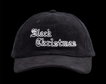 May include: Black corduroy baseball cap with the text "Black Christmas" embroidered in white gothic lettering. The cap has a curved brim and a small button on top. The background is solid black.