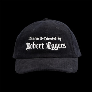 以下が含まれることがあります： 黒色のコーデュロイ野球帽。白のゴシック体で「Written & Directed by Robert Eggers」の文字が刺繍されています。つばはカーブしており、上部に小さなボタンが付いています。背景は黒色です。