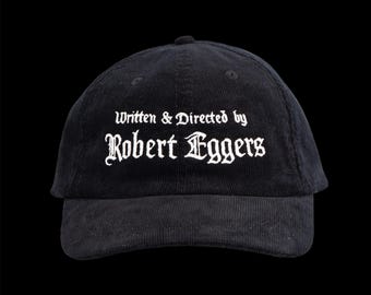 May include: Black corduroy baseball cap with the text "Written & Directed by Robert Eggers" in white gothic lettering. The cap has a curved brim and a small button on top. The background is solid black.