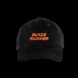Puede incluir: Una gorra de béisbol de pana negra con el texto BLADE RUNNER bordado en hilo naranja brillante en la parte frontal central. La gorra tiene una corona estructurada, una visera curva con costuras visibles y un botón en la parte superior.