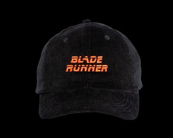 May include: A black corduroy baseball cap featuring the text BLADE RUNNER embroidered in bright orange thread across the front center. The cap has a structured crown, a curved brim with visible stitching, and a button on the top center.
