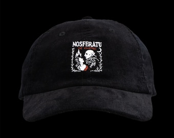 May include: Black corduroy baseball cap with the word "NOSFERATU" embroidered in white above a graphic of a vampire and a woman. The cap has a curved brim and a fabric strap closure.