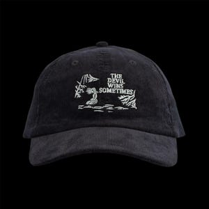 May include: Black corduroy baseball cap with white embroidered text reading "THE DEVIL WINS SOMETIMES". The cap has a curved peak and a structured crown. The design includes a small illustration.