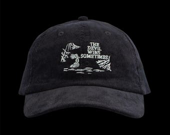 May include: Black corduroy baseball cap with the embroidered text "THE DEVIL WINS SOMETIMES" in white. The cap features a curved brim and a structured crown. The design includes a small illustration.