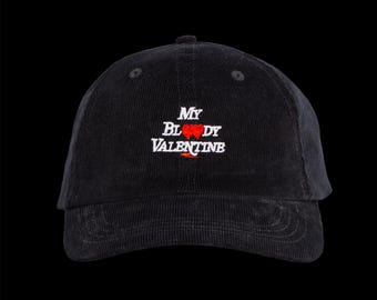 Peut inclure: Casquette de baseball en velours côtelé noir avec l'inscription "My Bloody Valentine" brodée en blanc et rouge. La casquette a une visière incurvée et un bouton sur le dessus. Le fond est noir.