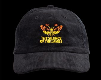 May include: Black corduroy baseball cap with an embroidered moth design in orange and yellow. The text "THE SILENCE OF THE LAMBS" is below the moth. The cap has a curved brim and a top button.