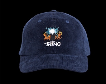 May include: Navy blue corduroy baseball cap with an embroidered design. The design features a stylized image with orange and white accents, and the word "THE THING" in white.