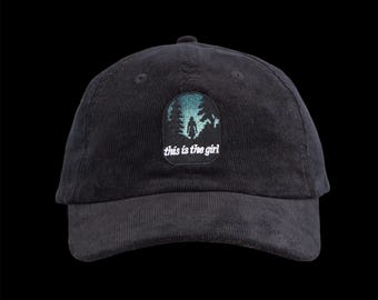May include: Black corduroy baseball cap with a curved brim. The cap features an embroidered patch with a forest scene and the text "this is the girl" in white. The cap has a button on top and an adjustable strap.