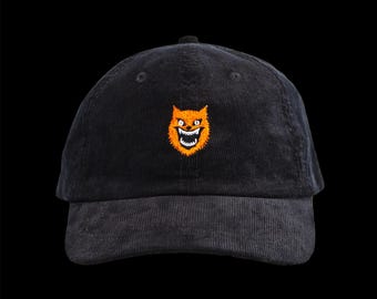 May include: Black corduroy baseball cap with an embroidered orange and white grinning cat face on the front. The cap has a curved brim and an adjustable strap at the back. The hat is a classic, casual accessory.
