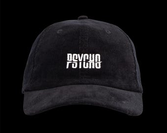 May include: Black corduroy baseball cap with the word "PSYCHO" embroidered in white across the front. The text has a stylized, distressed design. The cap has a curved brim and a button on top.