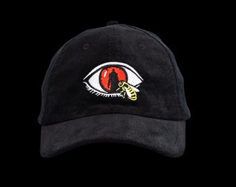 May include: Black corduroy baseball cap featuring an embroidered eye design with a red iris and a yellow and black bee. The eye design has a silhouette of a person inside. The cap has a curved brim and an adjustable strap.