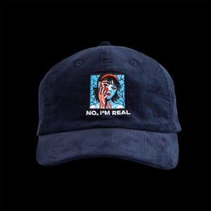 May include: Navy blue corduroy baseball cap with a square embroidered patch featuring a distressed illustration of a person with red accents and the text "NO, I'M REAL". The cap has a curved brim and a top button.