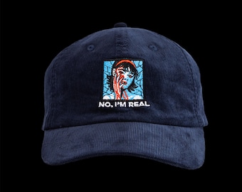 May include: Navy blue corduroy baseball cap with a square embroidered patch featuring a distressed illustration of a person with red accents and the text "NO, I'M REAL". The cap has a curved brim and a top button.