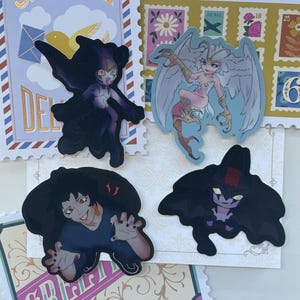 Op de afbeelding: Vier stickers met fantasiekarakters. Eén sticker toont een donkergekleurd wezen met vleermuisachtige vleugels. Een andere toont een gevleugelde humanoïde met een bleke huid en een vogelachtig been. De andere twee tonen donkerharige karakters.