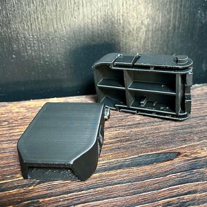 May include: A black, hinged, rectangular container with an open compartment and shelves. The lid is detached and lies next to the open container. The container is made of a matte black material and sits on a wooden surface.