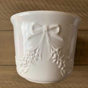 May include: A white ceramic planter with a glossy finish. The planter features a raised bow and floral design. The design is repeated around the base of the planter. The planter is sitting on a wooden surface.
