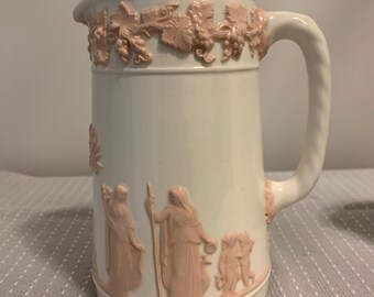May include: White ceramic pitcher with a pink relief design. The pitcher features a handle and a spout. The pink relief includes grape vines and classical figures. A decorative piece for serving beverages.