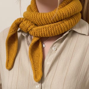 Puede incluir: Bufanda de punto amarillo mostaza. La bufanda está envuelta alrededor del cuello y atada por delante. La bufanda está hecha de un hilo grueso y texturizado. La persona lleva una camisa abotonada de color beige claro.