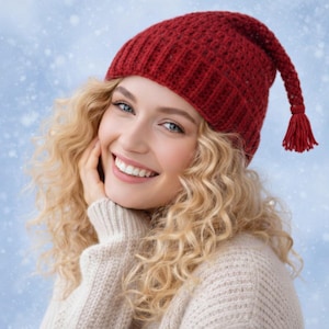 Puede incluir: Un gorro rojo tejido con una larga borla. El gorro está hecho de un material texturizado y cálido. La persona lleva un suéter color crema. El fondo es azul claro con copos de nieve blancos.