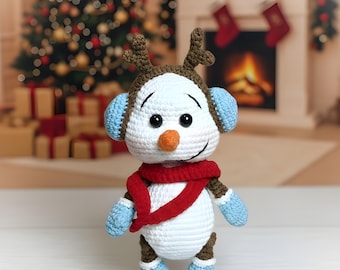 May include: A handmade crochet snowman doll with brown antlers, blue earmuffs, and a red scarf. The snowman has a carrot nose, black eyes, and blue mittens and boots with snowflake details. The background features a Christmas tree and fireplace.