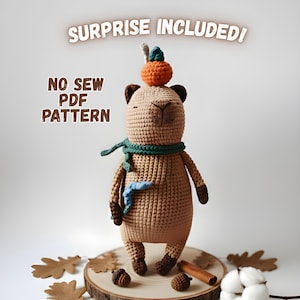 Peut inclure: Une poupée ours au crochet avec un chapeau citrouille et une écharpe verte. L'ours est beige avec des détails marron et un accent bleu. Le texte "SURPRISE INCLUDED!" et "NO SEW PDF PATTERN" sont visibles.