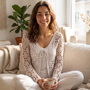 Könnte beinhalten: Eine langärmelige, cremeweiße Häkelstrickjacke mit einem komplizierten, blumenartigen Lochmuster. Das Kleidungsstück hat einen V-Ausschnitt und einen zarten Bindegürtel in der vorderen Mitte und wird über einem schlichten, einfarbigen weißen Oberteil getragen.