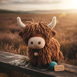 May include: A handmade crochet Highland cow with brown fur, beige horns, and a light beige muzzle. The cow is sitting on a wooden surface with a ball of teal yarn and a tag that says "Handmade with Love".