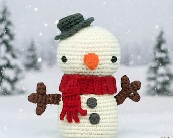 May include: A crocheted snowman with a white body, wearing a red scarf, a gray hat, and brown stick arms. The snowman has an orange carrot nose and black button eyes. The background shows a snowy winter scene with pine trees.