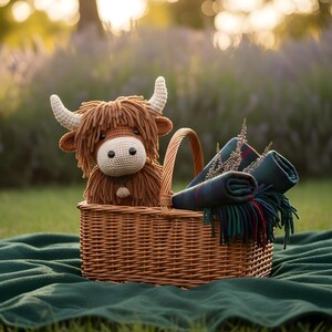 May include: A woven wicker picnic basket holds a crocheted Highland cow plush toy with brown fur and cream-colored horns. Rolled plaid blankets with fringe and sprigs of greenery complete the scene, set on a green blanket.