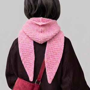 Puede incluir: Una bufanda con capucha de color rosa claro en tejido de punto gofrado con extremos largos y puntiagudos. La bufanda presenta un patrón de rejilla texturizado y se lleva sobre una chaqueta marrón oscuro con un bolso bandolera rojo oscuro.
