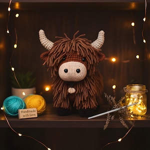 May include: A handmade crochet Highland cow with brown fur, beige face, and curved horns. The cow is displayed on a shelf with yarn, a jar of fairy lights, and a sign that says "Handmade with Love".