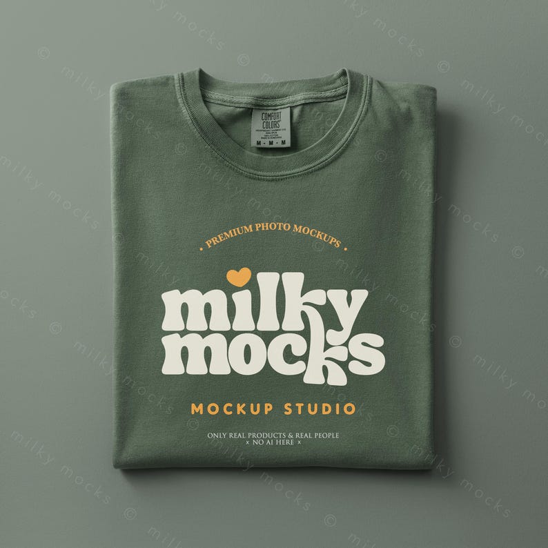 May include: Folded olive green t-shirt with the text "milky mocks" in a retro font, a small orange heart, and the words "MOCKUP STUDIO". The shirt has a tag that says "COMFORT COLORS".