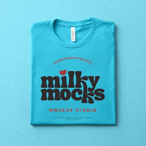 Puede incluir: Una camiseta azul claro doblada con cuello redondo. La camiseta presenta el texto "milky mocks" en una fuente marr&oacute;n retro, con un coraz&oacute;n rojo encima de la palabra. El texto adicional incluye "PREMIUM PHOTO MOCKUPS" y "MOCKUP STUDIO".