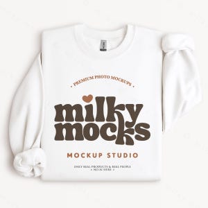K&ouml;nnte beinhalten: Ein wei&szlig;es Sweatshirt mit Rundhalsausschnitt und dem braunen Text "milky mocks", "PREMIUM PHOTO MOCKUPS" und "MOCKUP STUDIO". Die &Auml;rmel sind hochgekrempelt. Das Etikett lautet "GILDAN".