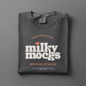 May include: A folded charcoal gray t-shirt with a crew neck. The shirt features the text "milky mocks" in a retro font, with a small orange heart above the word. Below the text is "MOCKUP STUDIO" and "ONLY REAL PRODUCTS & REAL PEOPLE NO AI HERE".