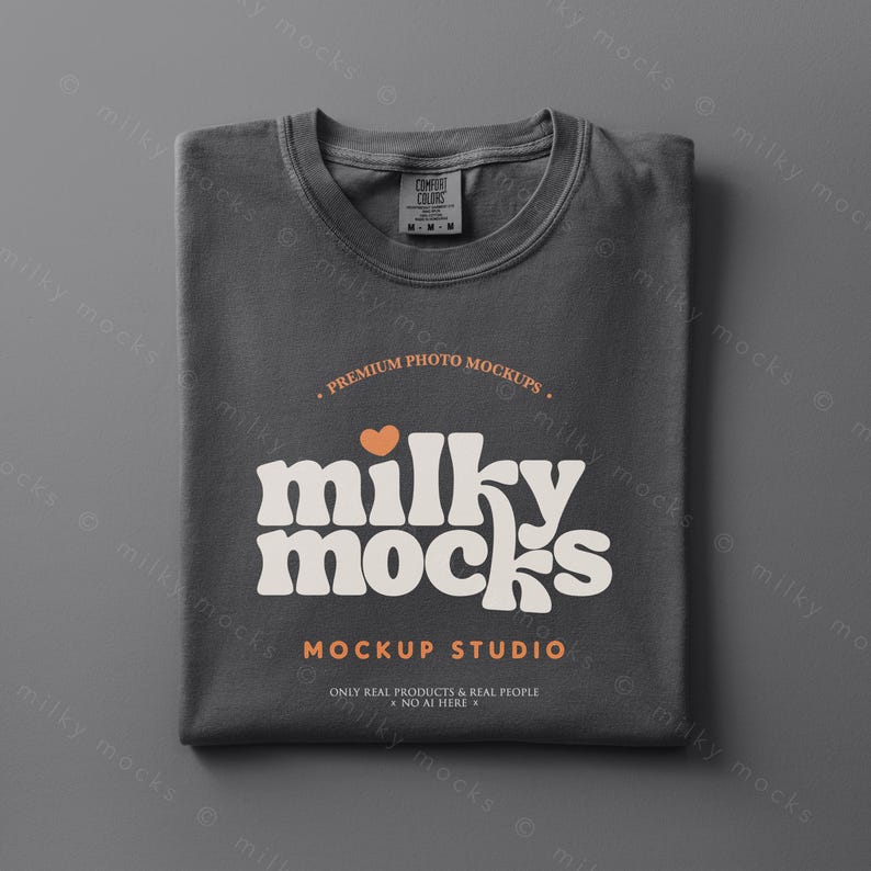 May include: A folded charcoal gray t-shirt with a crew neck. The shirt features the text "milky mocks" in a retro font, with a small orange heart above the word. Below the text is "MOCKUP STUDIO" and "ONLY REAL PRODUCTS & REAL PEOPLE NO AI HERE".