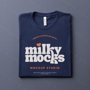 Puede incluir: Una camiseta azul marino doblada con cuello redondo. La camiseta muestra el texto "milky mocks" en un estilo retro, con un peque&ntilde;o coraz&oacute;n naranja encima. El texto adicional dice "PREMIUM PHOTO MOCKUPS" y "MOCKUP STUDIO".